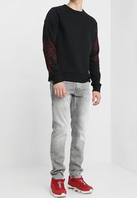 Svart sweatshirt med röd blommig broderi på ärmarna, tillsammans med ljusgrå jeans och röda sneakers med vita detaljer.
