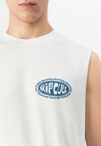 Közelkép egy személyről, aki fehér ujjatlan pólót visel, kék ovális Rip Curl logóval és "Raw Energy" felirattal a mellkasán.