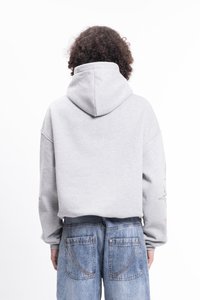 Szary hoodie z kangurzą kieszenią, w oversize'owym kroju, ze sznurkiem przy kapturze. Noszony z jasnoniebieskimi dżinsami, które mają dwie tylne kieszenie.
