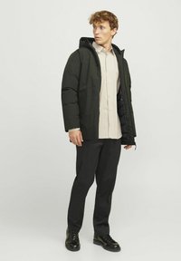 Chaqueta acolchada negra con capucha, que presenta un cierre frontal y bolsillos laterales. Llevada sobre una camisa de rayas claras y pantalones negros, con zapatos negros.