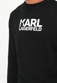 Melnā sporta krekla izgatavots no mīksta auduma ar baltu tekstu "KARL LAGERFELD" un stilizētu logotipu. Ar apaļu kakla izgriezumu, garām piedurknēm.