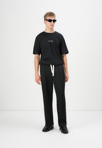 Black t-shirt with printed logo, loose fit. Black trousers with white drawstring, straight cut. Model wearing black sunglasses and shoes.