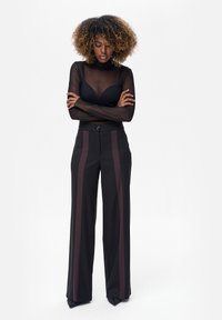 Kobieta o kręconych włosach ubrana w przezroczystą czarną bluzkę z długim rękawem, na czarnym biustonoszu oraz czarne, wysokie spodnie z ciemnymi pionowymi paskami.