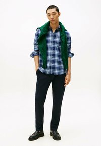 Camicia a quadri blu e verde, pantaloni blu scuro, maglione a trecce verde drappeggiato sulle spalle, scarpe nere, abbigliamento casual, vestibilità moderna.
