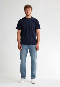 Marineblauw katoenen T-shirt met een ronde hals, korte mouwen, gecombineerd met lichtblauwe jeans en witte sneakers. Model staat tegen een effen achtergrond.