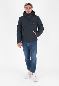 Ciemnoniebieska kurtka ocieplana z kapturem, zapięciem na zamek z przodu i bocznymi kieszeniami. Posiada małą naszywkę z logotypem na piersi. Noszona z niebieskimi dżinsami i białymi trampkami.