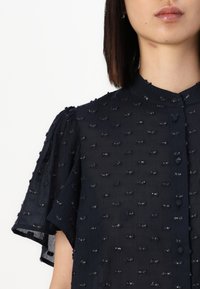 Blusa trasparente blu navy con maniche corte a palloncino, tessuto testurizzato caratterizzato da piccole imbottiture in rilievo e patta frontale con bottoni.