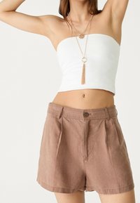 Top bianco senza spalline e pantaloni a vita alta marroni con pieghe. I pantaloni presentano una chiusura con bottone e tasche laterali.