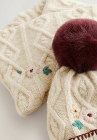 Strik en cremet hat med kabelmønstre, der har en burgunderrød faux fur pom-pom og broderede blomster i pink, gul og grønne accenter.