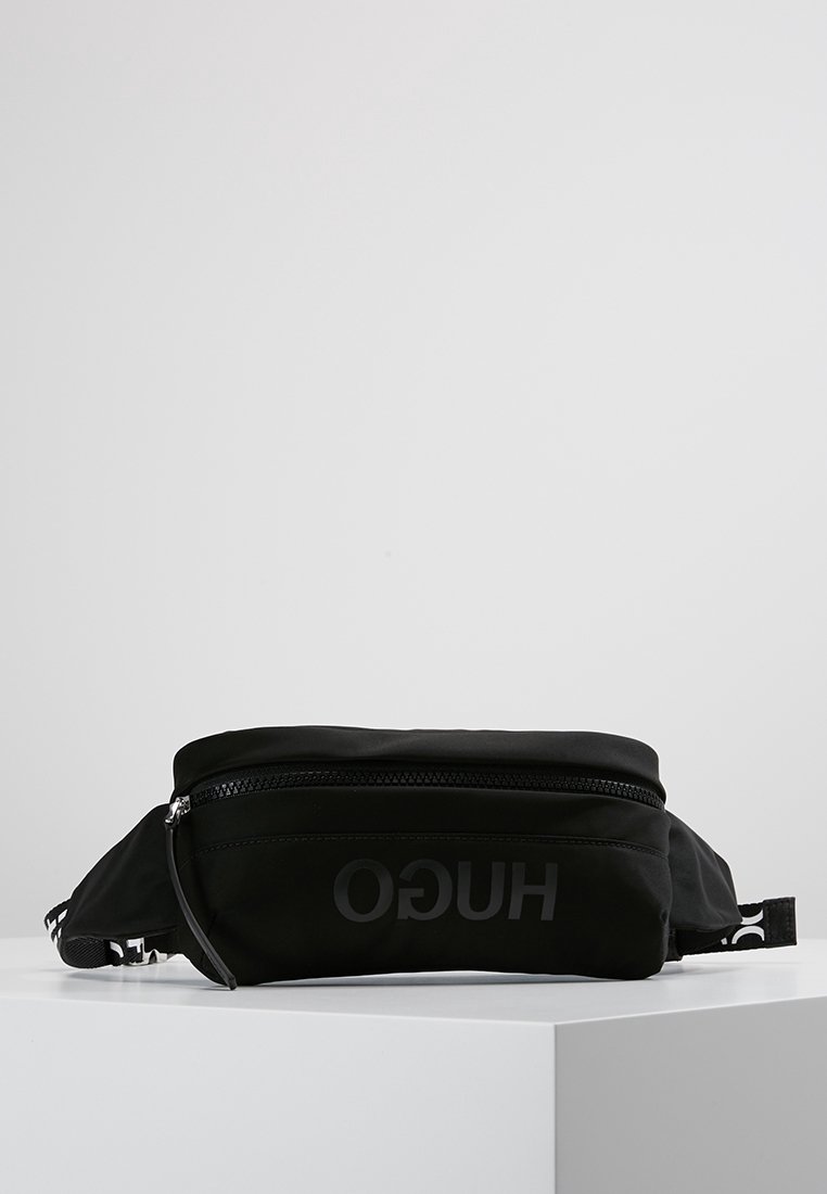 Sac banane noir de marque HUGO avec une poche zippée à l'avant, présenté sur une surface blanche sur un fond gris clair uni.