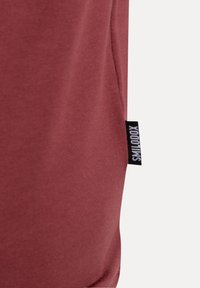 Närbild av vinr�öd tyg med en liten svart etikett med vit text, sydd i sidogömman.