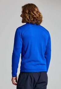 Uomo con capelli ricci a lunghezza spalle, che indossa una camicia a maniche lunghe blu brillante e pantaloni neri, girato di spalle contro uno sfondo semplice.