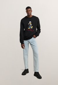 Fekete pulóver színes Donald Duck grafikával és piros felirattal. A modell világoskék farmernadrágot és fekete sportcipőt visel. Simább textúra, laza illeszkedés.