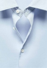 Chemise bleu clair à boutons avec un tissu texturé. Les caractéristiques notables incluent un col classique et deux boutons à l'ouverture du cou.