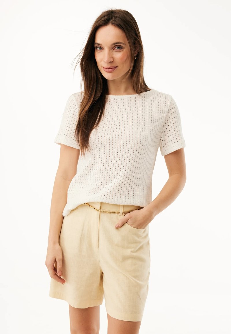 Vrouw met lang bruin haar, gekleed in een wit gebreid topje met korte mouwen en beige shorts met hoge taille en een gouden kettingriem, staande met één hand in haar zak.