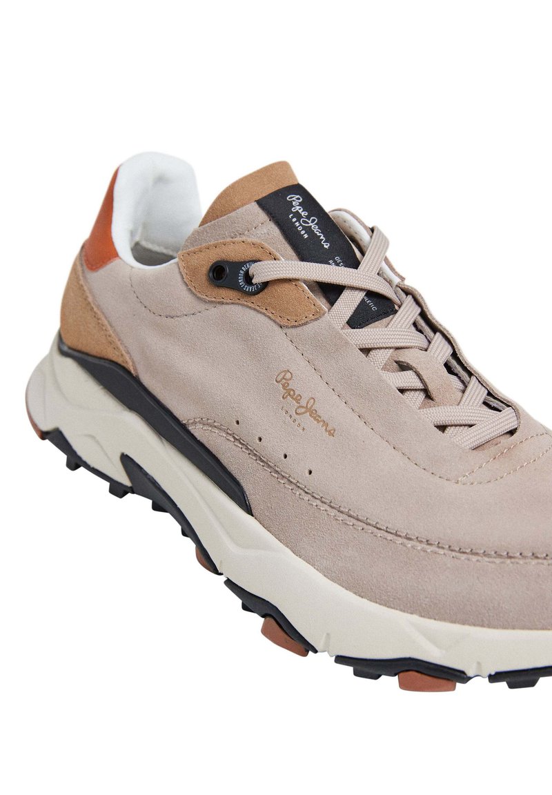 Zapatillas de ante beige con acentos en marrón y negro, suela intermedia blanca gruesa, cordones beige y una lengüeta de talón negra texturizada con un logo.