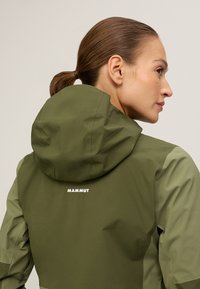 Groene outdoorjas met capuchon, met een slank ontwerp, een gladde textuur en de merknaam "MAMMUT" gedrukt op de achterkant.