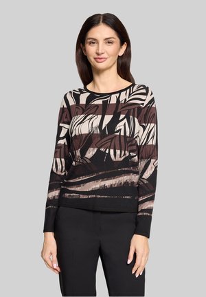 Frau mit dunklem, glattem Haar trägt ein langärmliges Oberteil mit abstraktem Muster in Schwarz, Beige und Braun sowie eine schwarze Hose und steht vor einem grauen Hintergrund.