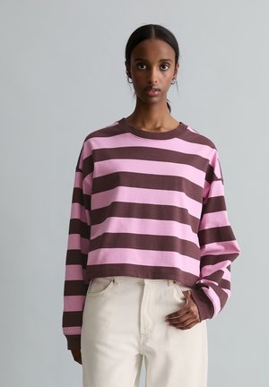 Young woman with dark hair wears oversized pink and brown striped long-sleeve shirt and light beige pants, standing against plain background.