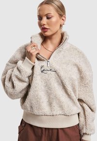 Fuzzy cream pullover with a half-zip collar, ribbed hem, and cuffs; features a subtle embroidered logo on the front. Soft, textured fabric.