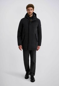 Man in een zwarte winterjas met capuchon, donkere broek, zwarte nette schoenen, staande tegen een effen lichte achtergrond.