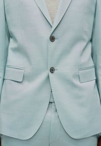 Chaqueta de traje azul claro a medida con dos botones oscuros abrochados sobre pantalones a juego, mostrando la parte media frontal y los bolsillos.