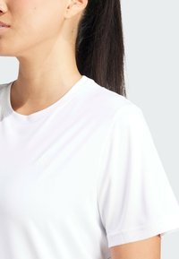 Valkoinen urheilupaita lyhyillä hihoilla ja pyöreällä kaula-aukolla, jossa on hienovarainen kohotettu logo rinnassa ja sileä pinta.