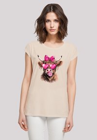 Beige cotton T-shirt featuring a graphic of a cow with a pink bow, heart-shaped sunglasses, and a lollipop, accented with detailed fur illustration.