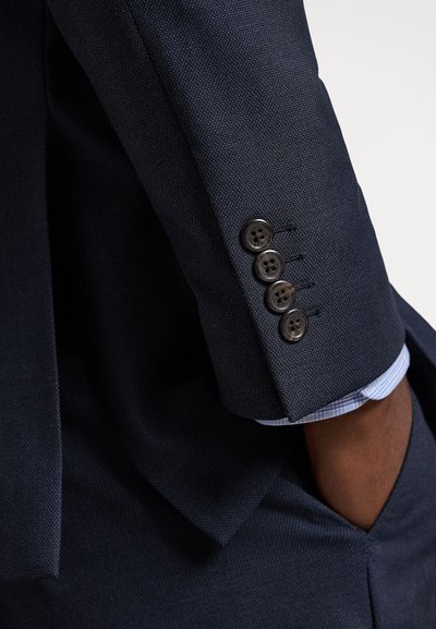 Tumši zila uzvalka piedurkne ar četrām tumšām pogām, redzams gaiši zilas rūtainas krekla aproces gals, un roka ievietota atbilstoša tumsā zilā kabatā.