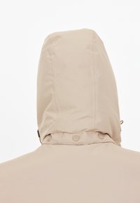 Beige huppari, jossa on teksturoitu kangas ja säädettävät nyörit. Hupussa on painonapit pohjassa varmistaen tiukan istuvuuden.