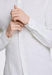 Vit linneskjorta med knäppning framtill, synliga knappar och uppvikta ärmar. Tyget har en lätt texturerad yta. Händerna är placerade nära kragen.