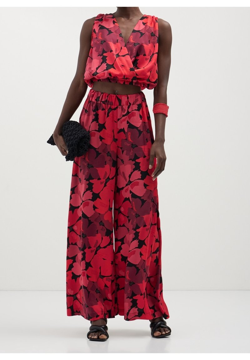 Femme portant un crop top sans manches assorti rouge et noir à motifs floraux et un pantalon large, tenant une pochette noire tissée et portant des sandales noires.