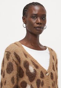 Brun leopardmønstret cardigan laget av mykt fleece-materiale med dyp V-hals og knappelukking, brukt over en hvit topp.