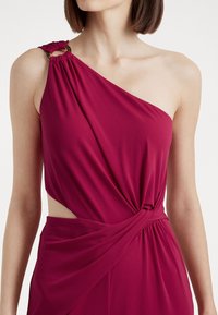 Fuksiin värinen hihaton mekko, jossa on kiertävä muotoilu, sivuaukko ja ryppyinen olkaimen osa, jossa on pyöreä kultasävyinen koriste.