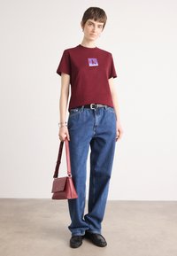 Burgunderfarget bomulls t-skjorte med en lilla logo, kombinert med blå jeans, svarte sko og en rød crossover-veske. Glatte teksturer og avslappet passform.