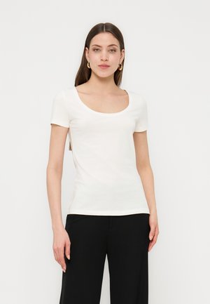 Femeie cu păr lung și întunecat, purtând cercei aurii în formă de cerc, un top alb cu mâneci scurte și decolteu rotund și pantaloni negri, stând în picioare pe un fundal alb.