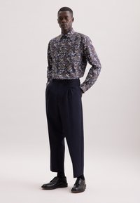 Camicia blu con motivo floreale e maniche lunghe, abbinata a pantaloni blu a vita alta e plissettati e scarpe eleganti nere. Texture liscia.