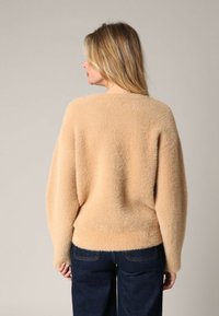 Vrouw met schouderlang blond haar, gekleed in een beige pluizige trui en donkerblauwe jeans, met haar rug naar de camera gekeerd tegen een effen achtergrond.