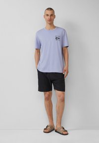 Lichtpaars, kortom mouw T-shirt met zwart grafisch ontwerp, gecombineerd met zwarte shorts en bruine sandalen, op een neutrale achtergrond.