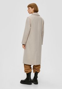 Beige Wollmischungsmantel mit geradem Schnitt, Reverskragen und seitlichen Taschen, getragen über braunen Hosen mit Aufschlag und schwarzen Stiefeletten.