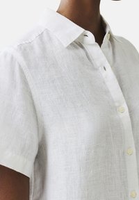 Camicia di lino bianca con maniche corte, colletto standard e chiusura frontale con bottoni. Tessuto testurizzato con un aspetto leggermente trasparente.