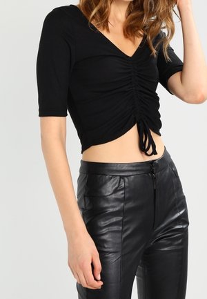 Femme portant un crop top noir froncé à manches courtes et un pantalon en cuir noir taille haute sur un fond uni.