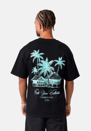 Mand med flettet hår iført en sort T-shirt med blå palmer og hytter med teksten "Sixth June Collection Caribbean Soul" på ryggen.