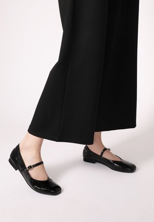 Black patent leather Mary Jane shoes with a strap and small buckle detail. Flat sole with a rounded toe design. Pair with wide black trousers.