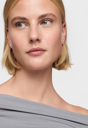 Joven con cabello corto y rubio lleva pendientes de bola rosa y una blusa gris con los hombros descubiertos, mirando ligeramente hacia la izquierda sobre un fondo blanco.