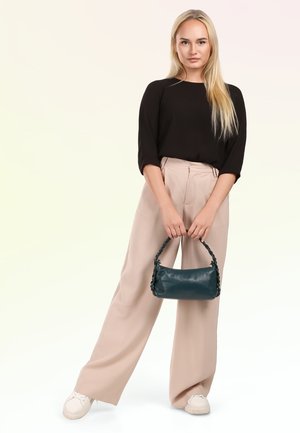 Blonde vrouw in een zwarte top en beige broek met wijd uitlopende pijpen houdt een kleine donkergroene handtas vast en staat met één voet iets naar voren.