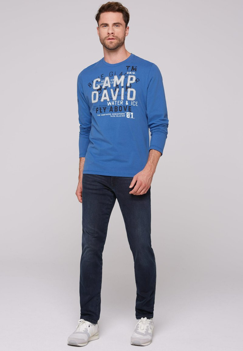 Niebieska koszulka z długim rękawem z graficznym napisem, połączona z ciemnymi dżinsami i jasnymi trampkami. Koszulka ma okrągły dekolt i miękką fakturę.