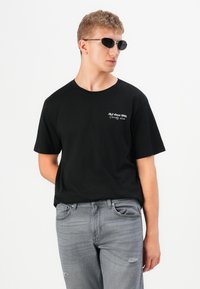 Joven con gafas de sol negras, camiseta negra con texto blanco y jeans grises desvanecidos, de pie contra un fondo blanco.