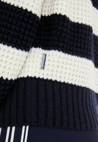 Strickpullover mit navyblauen und cremefarbenen horizontalen Streifen, strukturiertem Stoff, gerippten Bündchen und einem kleinen Etikett mit der Aufschrift "Marcon Sports."