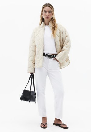 Vrouw met lang haar die een beige doorgestikte jas, een wit top, witte broek, zwarte riem, zwarte handtas en zwarte sandalen draagt, staand tegen een witte achtergrond.